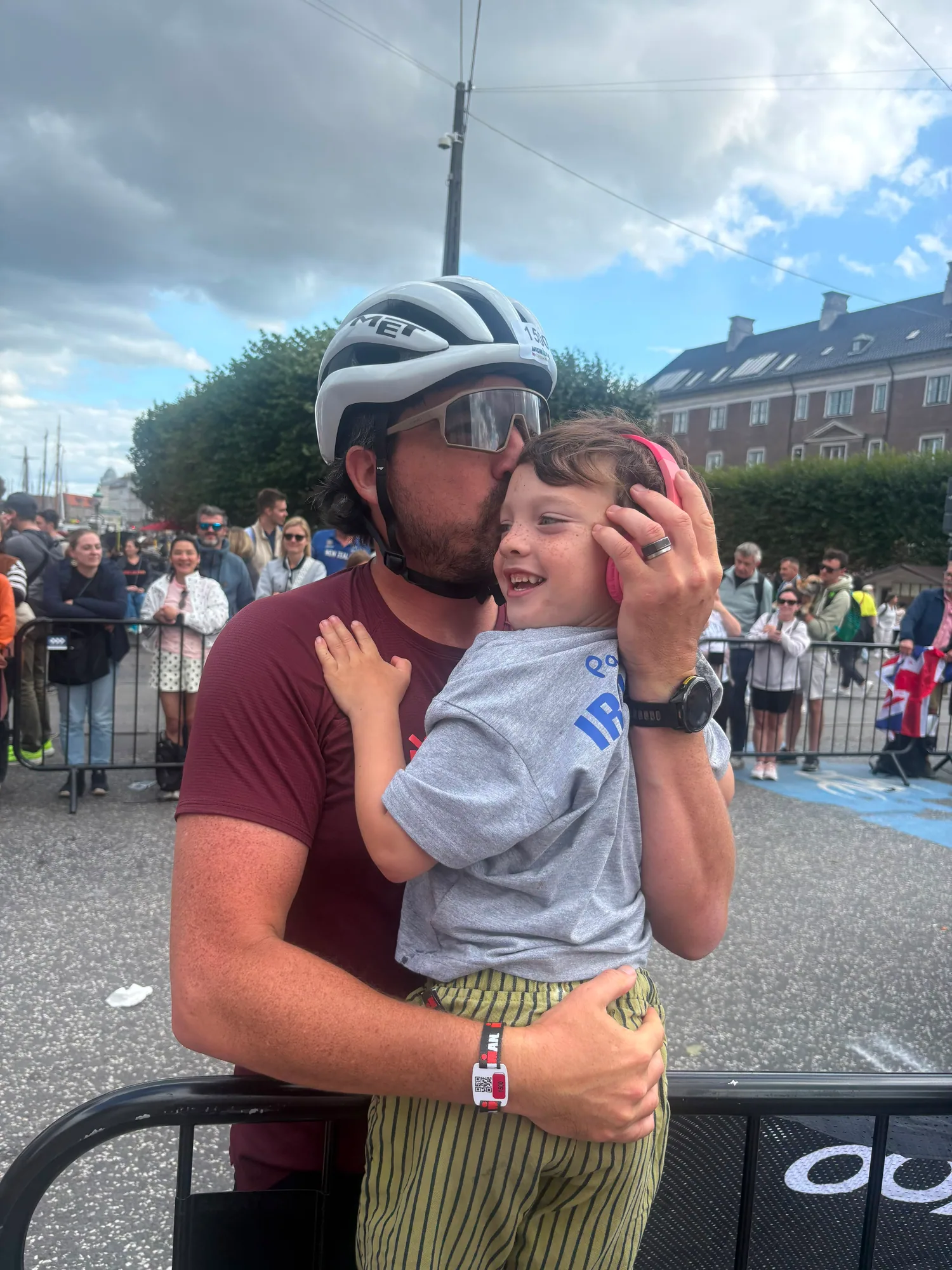 Bruno in his bike helmet kissing a smiling child wearing pink ear protectors beside the course barrier