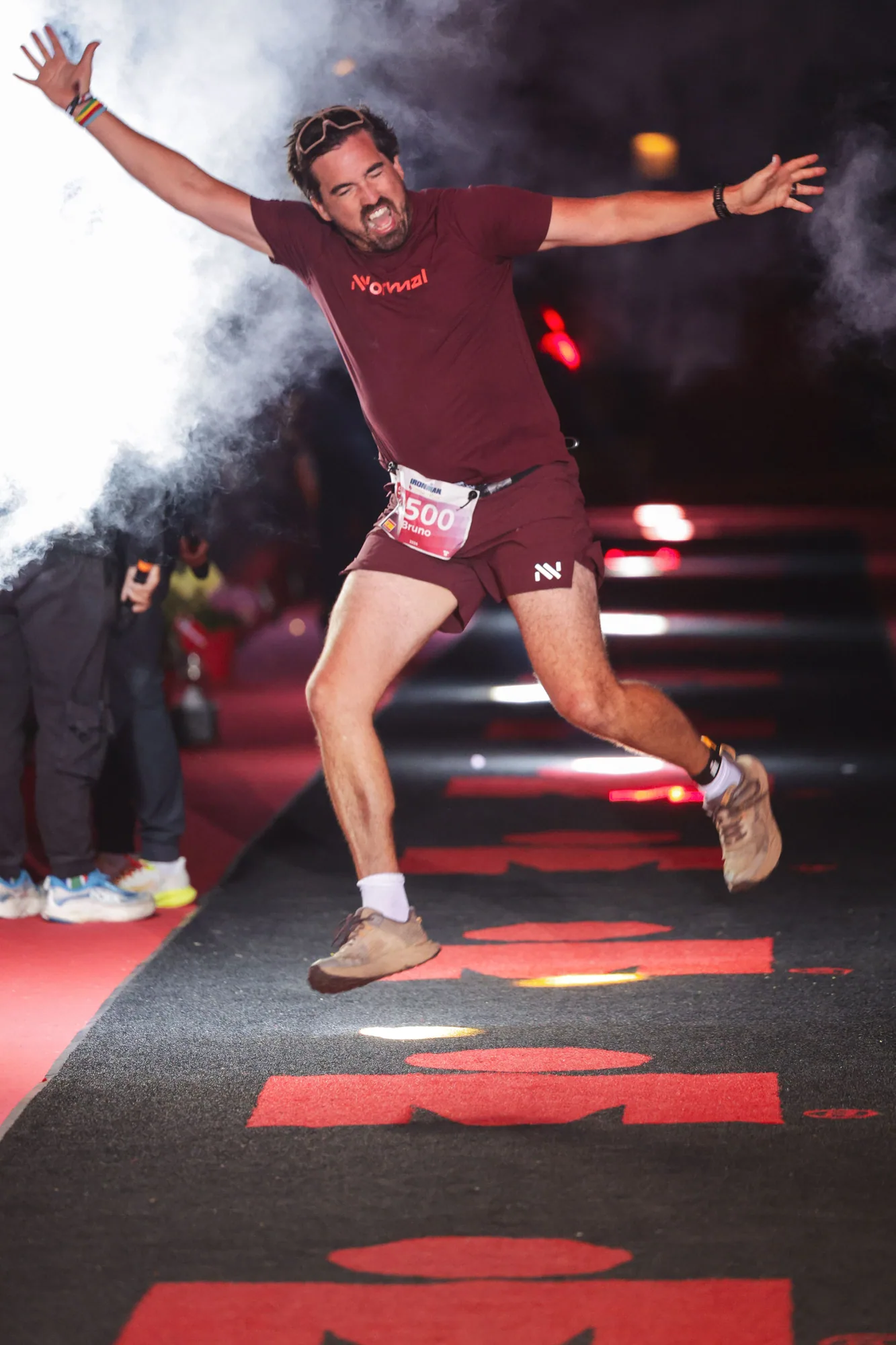 Bruno airborne with both arms spread above the red IRONMAN finish carpet amid white smoke