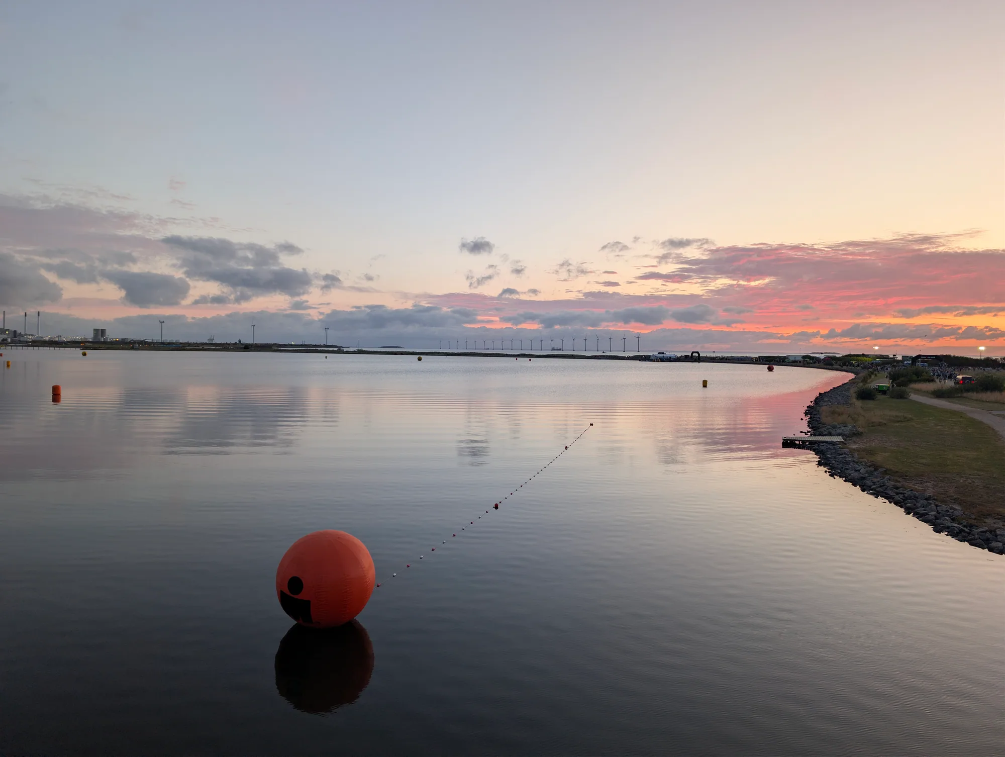 Calm swim lagoon before dawn reflecting a pink sky, with an orange IRONMAN buoy and wind turbines on the horizon
