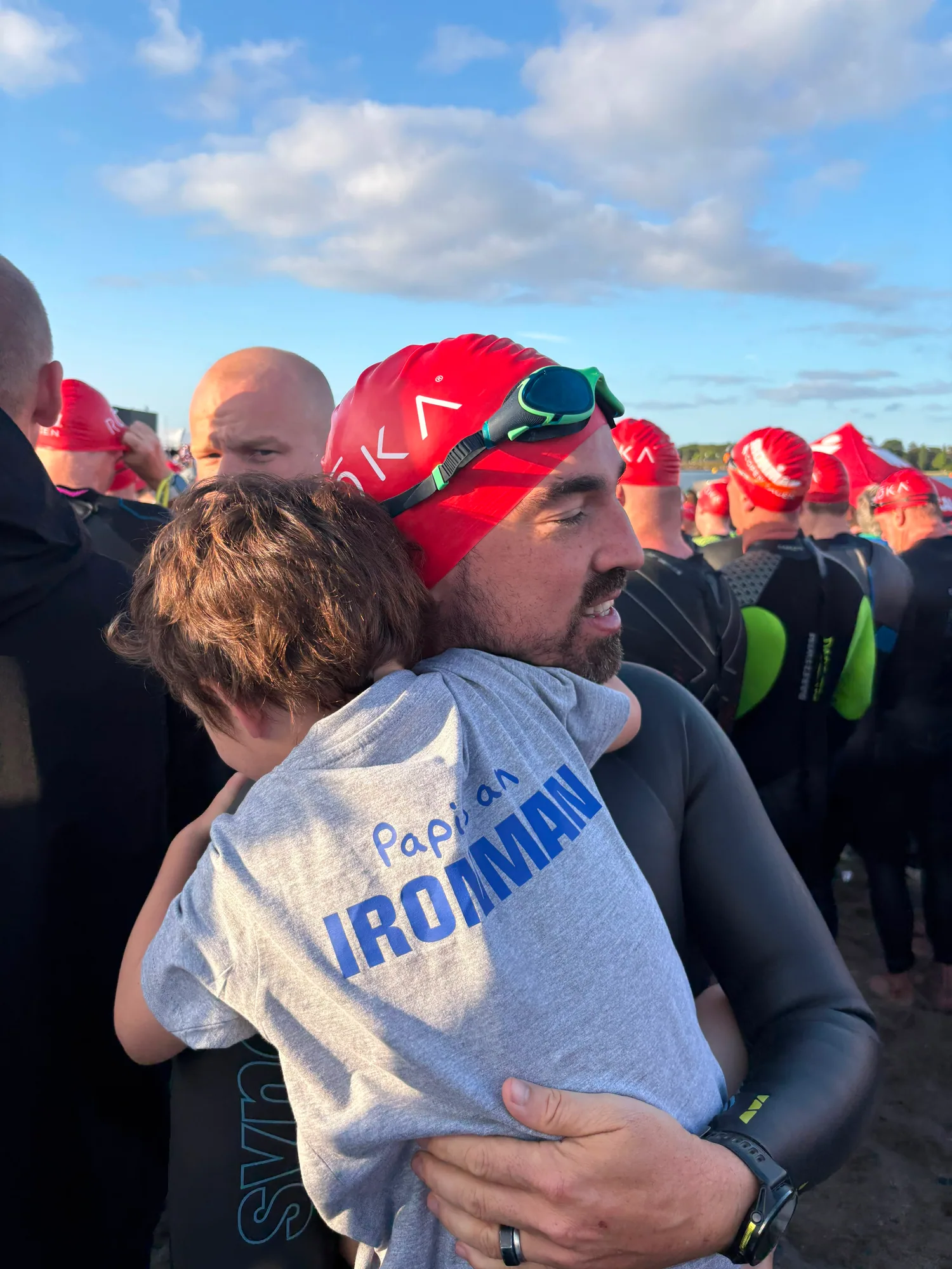 Bruno wearing a red swim cap and black wetsuit while hugging a child at the swim start